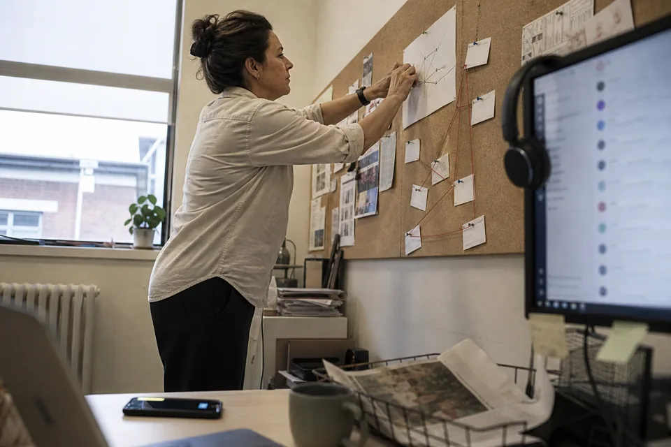 Communications coordinator at a desk pinning printed channel and inbox labels to a corkboard, a second monitor beside her showing a messaging app with a channel list out of focus