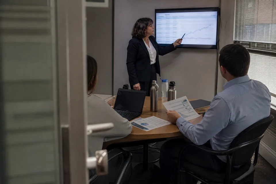 Head of communications pointing at a row on a wall screen while colleagues hold printed one-page reports
